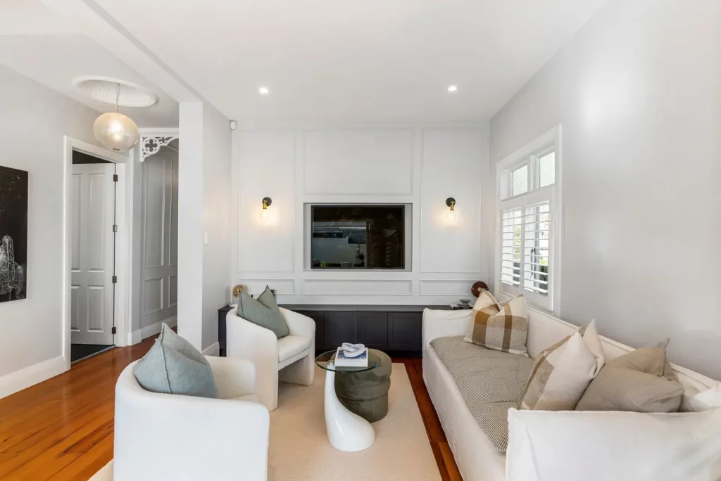 Modern living room with freshly painted white walls, wooden floors, a built-in television, a beige sofa with checked cushions, two white armchairs, a small glass coffee table, and large windows with plantation shutters letting in natural light. Ponsonby